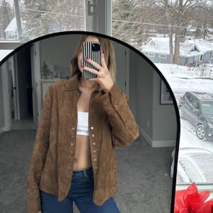 May include: A brown suede button-up shirt with long sleeves. The shirt has silver buttons down the front and on the cuffs. The person in the image is wearing blue jeans and a white top.