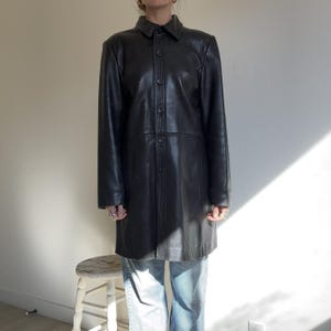 May include: A black leather coat with a button-up front and a collared neckline. The coat is knee-length and has long sleeves. The coat is worn over blue jeans. The coat is a classic style.
