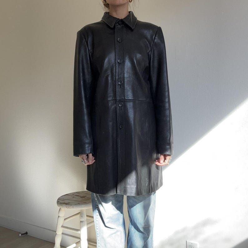 May include: A black leather coat with a button-up front and a collared neckline. The coat is knee-length and has long sleeves. The coat is worn over blue jeans. The coat is a classic style.