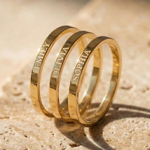 May include: Three gold rings, each engraved with a name: "EMILY", "OLIVIA", and "SOPHIA". The rings are stacked together. They are displayed on a textured, light-brown stone surface.