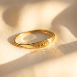 Op de afbeelding: Een goudkleurige ring met de naam "chloe" gegraveerd aan de voorkant. De ring ligt op een zachte, crèmekleurige stof. De ring heeft een gladde, gepolijste afwerking en een lichte kromming.