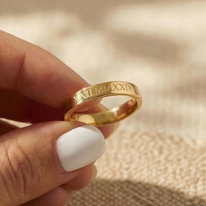 May include: A gold ring with Roman numerals engraved on the band. The ring is held up against a neutral background. The ring is a simple band design.