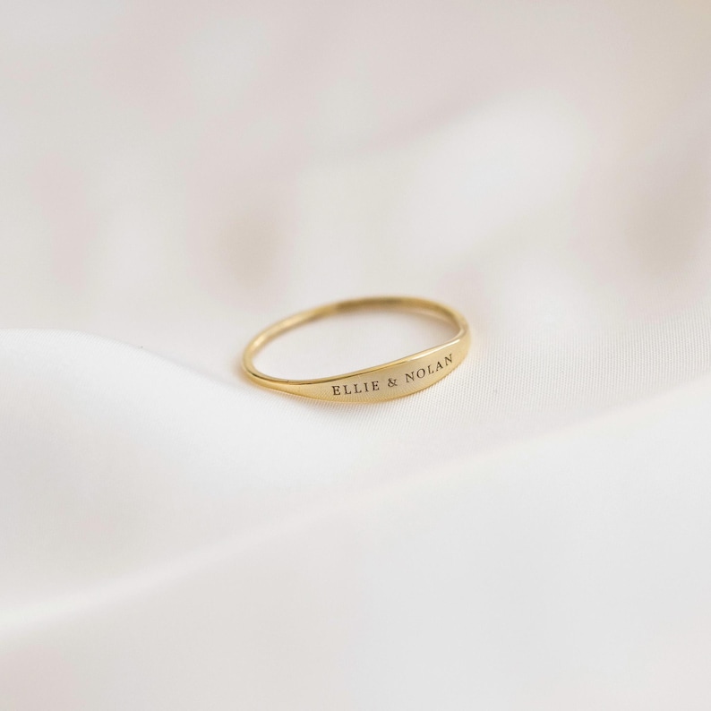 May include: A gold ring with a curved, rectangular design. The ring is engraved with the words 'ELLIE & NOLAN' in a simple, sans-serif font. The ring is set against a white, textured background, creating a minimalist aesthetic. The ring appears to be made of polished metal.