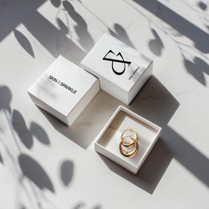 Puede incluir: Tres cajas blancas con el texto "SKIN & SPARKLE" y un logotipo. Una caja est&aacute; abierta, revelando pendientes de aro dorados. Los pendientes est&aacute;n en una caja blanca. La imagen est&aacute; iluminada con luz natural y sombras.