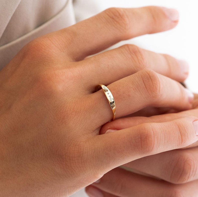 Puede incluir: Un anillo de oro con la inscripci&oacute;n "ELLA & NOL". El anillo se lleva en un dedo, mostrando su delicado dise&ntilde;o y acabado pulido. La inscripci&oacute;n del anillo a&ntilde;ade un toque personalizado a esta joya.