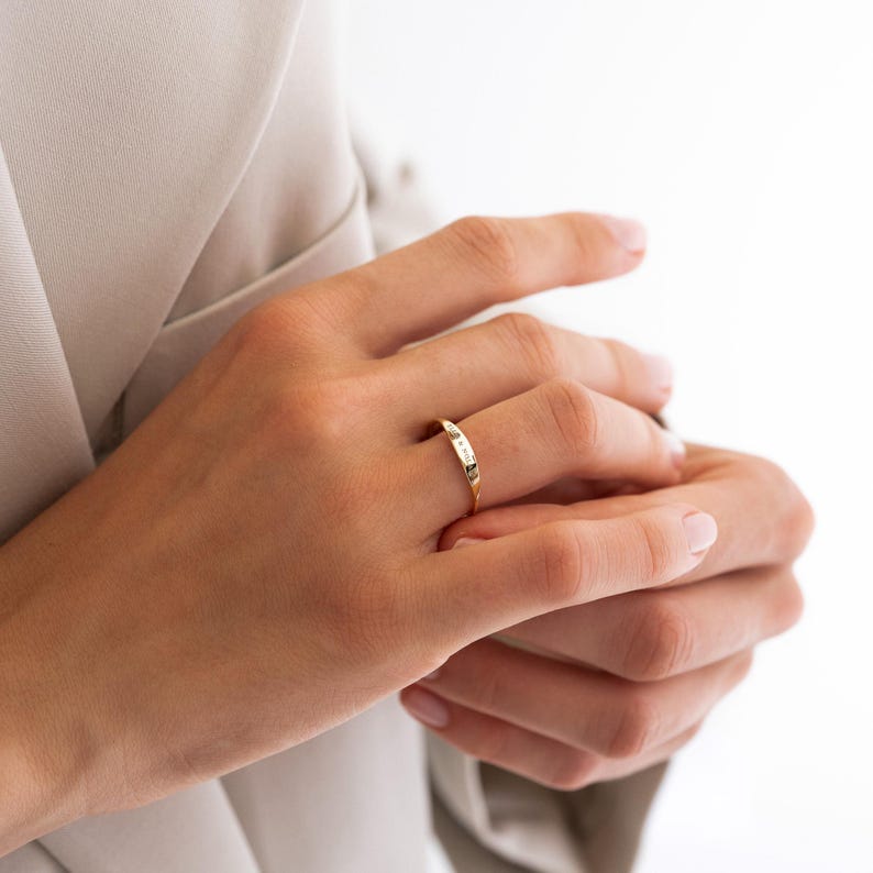 Puede incluir: Un anillo de oro con una cara rectangular adornada con peque&ntilde;as piedras claras. El anillo se lleva en un dedo, sobre un fondo neutro. El dise&ntilde;o del anillo es sencillo y elegante, adecuado para el uso diario.