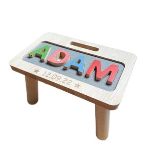 Puede incluir: Un taburete de madera clara con una superficie tipo rompecabezas que muestra el nombre ADAM en letras coloridas. Las letras verdes, rojas, azules y rojas están insertadas en un área gris. La fecha 12.09.22 está grabada entre dos estrellas en la madera.