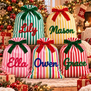 Pode incluir: Cinco sacos de presente de Natal em tecido com riscas verticais em verde, amarelo, rosa e vermelho. Cada saco apresenta um grande laço de veludo no topo e um nome personalizado em letra cursiva: Lily, Mason, Ella, Owen e Grace.