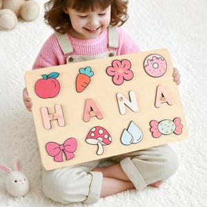 May include: A wooden puzzle board displaying the name "HANA" with colourful letters. The board includes shapes such as an apple, carrot, flower, donut, bow, mushroom, water drops, and candy. This puzzle is designed for children.