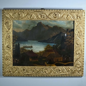 May include: A landscape painting in a rectangular gold frame with embossed thistle and leaf motifs. The artwork depicts a lake surrounded by dark mountains, with small buildings and trees in the foreground. The frame features a textured border.