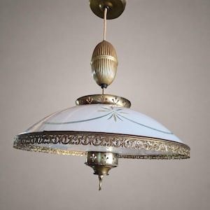 May include: A pendant light fixture featuring a white glass dome shade with a delicate gold-toned starburst pattern and a decorative brass filigree border. The fixture includes a ribbed brass counterweight and a central brass finial.