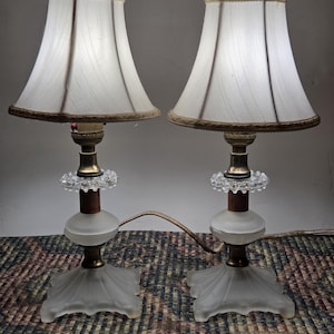 May include: Pair of vintage table lamps with white fabric shades, gold trim, and frosted glass bases. The lamps have a classic design with a decorative glass element and a brown wooden accent. The lamps are connected by a single cord.