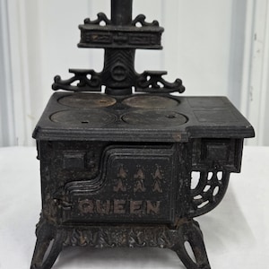 May include: A black cast iron toy stove with the word "QUEEN" on the front. The stove has four burners, a decorative back, and ornate legs. The stove is a miniature replica of a vintage kitchen appliance.