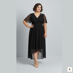 以下が含まれることがあります： Vネックと短いシアー袖の黒いミディドレス。ドレスは、フィットしたウエストと非対称のスカートが特徴です。肩には装飾が見えます。モデルは白いサンダルを履いています。