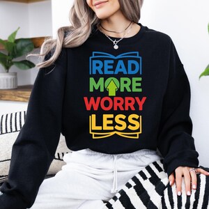 May include: Black sweatshirt with the text "READ MORE WORRY LESS" in blue, green, red, and yellow. The design includes an open book graphic above and below the text, and an upward-pointing green arrow.