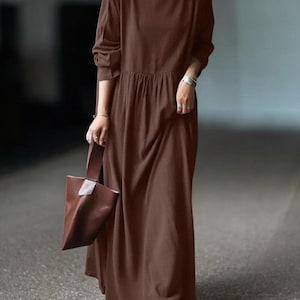 May include: A long, brown dress with long sleeves and a gathered waist. The dress is paired with a brown tote bag. The model is wearing gold earrings and a silver bracelet. The dress is made of a soft, flowing fabric.