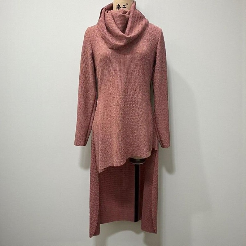 May include: A dusty rose-colored sweater dress with a cowl neck and long sleeves. The dress features a high-low hemline, with the back extending to a longer length. The fabric has a textured appearance.