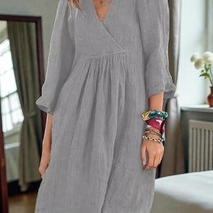 K&ouml;nnte beinhalten: Graues knielanges Kleid mit V-Ausschnitt und Dreiviertel&auml;rmeln. Das Kleid hat eine geraffte Taille und eine lockere Passform. Die &Auml;rmel haben eine leichte Glockenform. Der Stoff scheint eine leichte Leinen- oder Baumwollmischung zu sein.