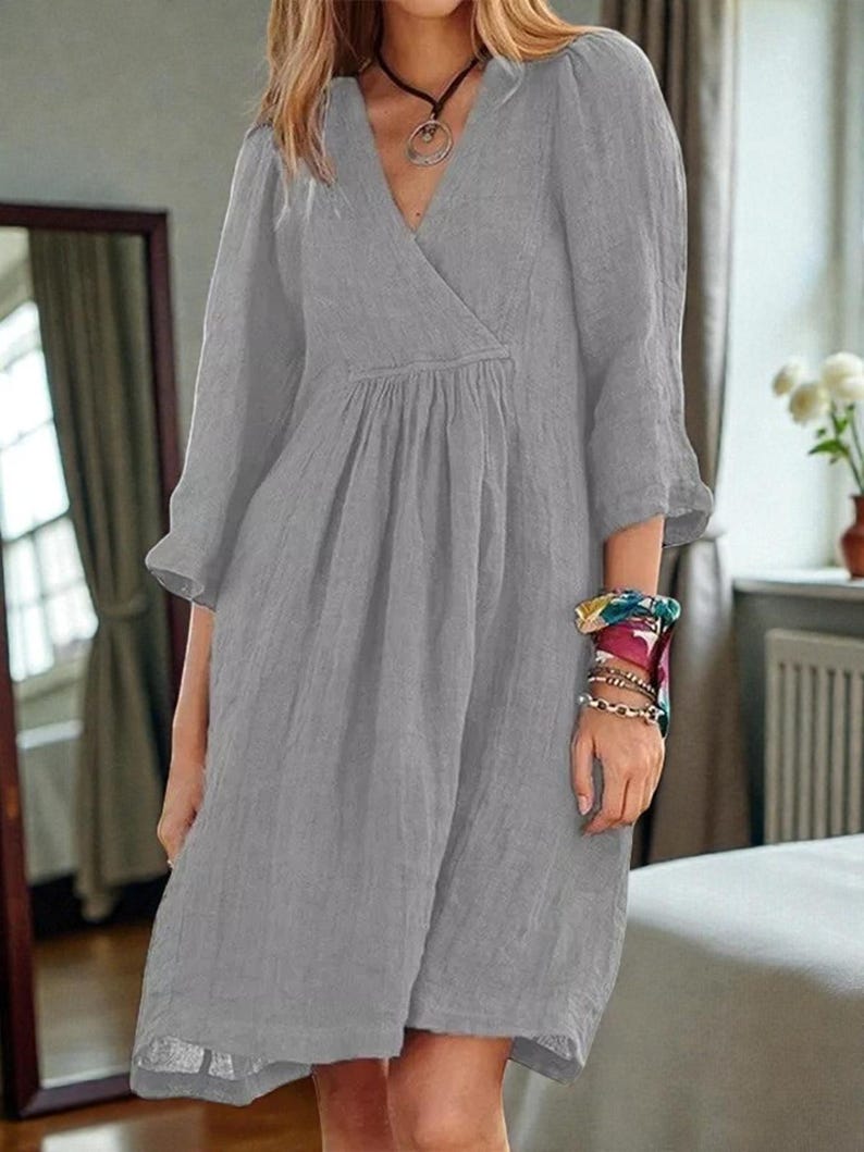 K&ouml;nnte beinhalten: Graues knielanges Kleid mit V-Ausschnitt und Dreiviertel&auml;rmeln. Das Kleid hat eine geraffte Taille und eine lockere Passform. Die &Auml;rmel haben eine leichte Glockenform. Der Stoff scheint eine leichte Leinen- oder Baumwollmischung zu sein.