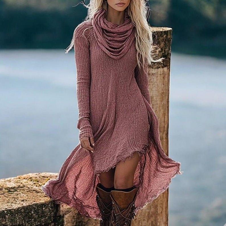 May include: A mauve-colored long-sleeve dress with a cowl neck and a high-low hemline. The dress is paired with brown leather boots. The model is standing outdoors near a body of water.