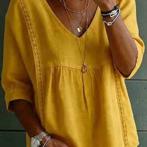 Könnte beinhalten: Senfgelbe V-Ausschnitt-Bluse mit kurzen Ärmeln. Die Bluse hat zarte Spitzentrimmungen am Ausschnitt und an den Ärmeln. Das Model trägt mehrere goldene Halsketten und Armbänder. Am linken Handgelenk ist eine silberne Uhr zu sehen.