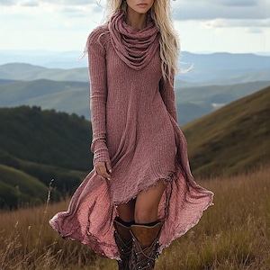 May include: A dusty rose-colored long-sleeve dress with a cowl neck and a high-low hemline. The dress is made of a ribbed knit fabric. The model is wearing brown leather boots. The background features rolling hills and a cloudy sky.