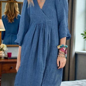 K&ouml;nnte beinhalten: Blaues Leinenkleid mit V-Ausschnitt und Dreiviertel&auml;rmeln. Das Kleid hat eine geraffte Taille und reicht bis zum Knie. Das Model tr&auml;gt eine Halskette und Armb&auml;nder. Das Kleid ist einfarbig und scheint aus leichtem Stoff zu sein.