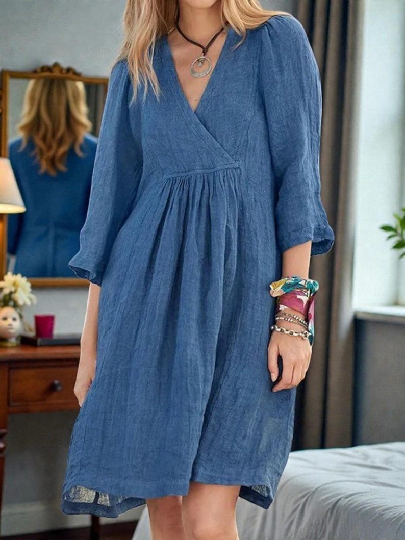 K&ouml;nnte beinhalten: Blaues Leinenkleid mit V-Ausschnitt und Dreiviertel&auml;rmeln. Das Kleid hat eine geraffte Taille und reicht bis zum Knie. Das Model tr&auml;gt eine Halskette und Armb&auml;nder. Das Kleid ist einfarbig und scheint aus leichtem Stoff zu sein.