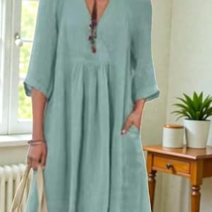 K&ouml;nnte beinhalten: Hellblaues Kleid mit V-Ausschnitt und Dreiviertel&auml;rmeln. Das Kleid hat Taschen und eine geraffte Taille. Eine Halskette mit Anh&auml;nger ist sichtbar. Das Kleid ist aus einem weichen, flie&szlig;enden Stoff gefertigt.
