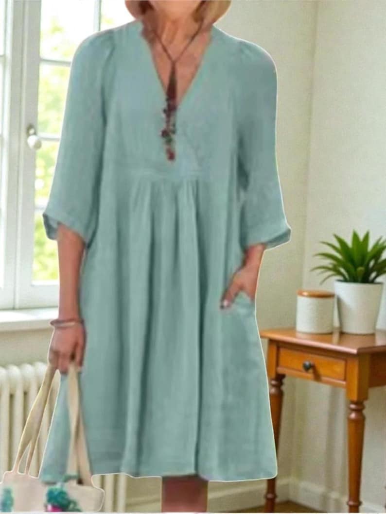 K&ouml;nnte beinhalten: Hellblaues Kleid mit V-Ausschnitt und Dreiviertel&auml;rmeln. Das Kleid hat Taschen und eine geraffte Taille. Eine Halskette mit Anh&auml;nger ist sichtbar. Das Kleid ist aus einem weichen, flie&szlig;enden Stoff gefertigt.