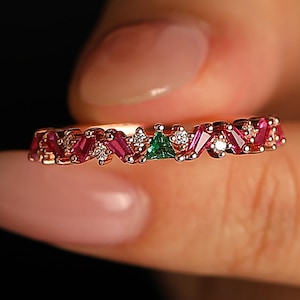 Może przedstawiać: Pierścionek z różowego złota z naprzemiennymi czerwonymi i białymi kamieniami szlachetnymi, z małym zielonym kamieniem szlachetnym i kilkoma małymi białymi kamieniami szlachetnymi. Pierścionek trzymany jest między palcami na ciemnym tle.