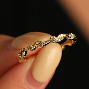 May include: A delicate gold ring featuring a repeating pattern of leaves and small, round gemstones. The ring is held between fingers, showcasing its intricate design and subtle sparkle. The ring's warm gold color contrasts with the neutral background.