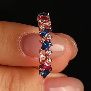 May include: A rose gold ring with a line of alternating blue, red, and clear gemstones. The gemstones are pear-shaped and round, set close together. The ring is held against a dark background.