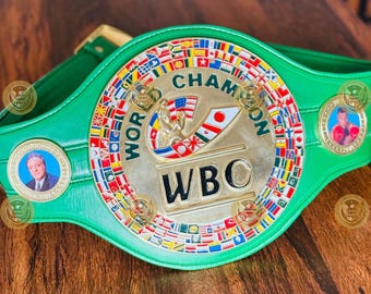 Op de afbeelding: Een groene lederen kampioenschapsriem voor boksen met een grote ronde goudkleurige middenplaat. De plaat toont de tekst WORLD CHAMPION en WBC, een centrale afbeelding van een bokser en een rand van kleine internationale vlaggen. Twee ronde goudkleurige zijmedaillons bevatten portretten van personen.