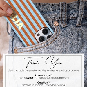May include: A phone case featuring a blue and orange striped design with the name "LIA" in white. The case is partially visible in a denim pocket. A white card with the text "Thank You" and shop details is also present in the image.