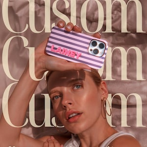 May include: A custom phone case with a pink and purple striped design, featuring the name "LAINEY" in pink letters. The case is held up in front of a person. The text "Your story, your style&mdash;encased in grace" is visible.