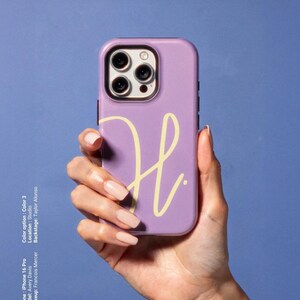 May include: A lavender iPhone 16 Pro case with a yellow cursive "H" design. The phone case is held in a hand with long, manicured nails. The background is a solid blue.