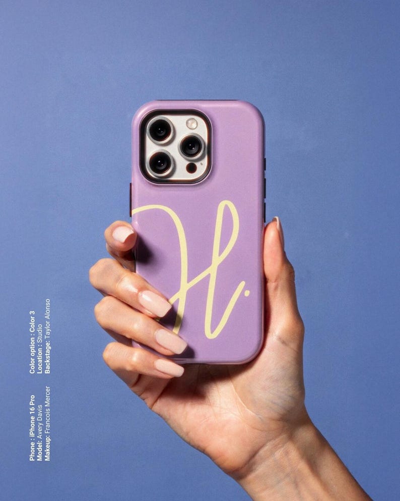 May include: A lavender iPhone 16 Pro case with a yellow cursive "H" design. The phone case is held in a hand with long, manicured nails. The background is a solid blue.