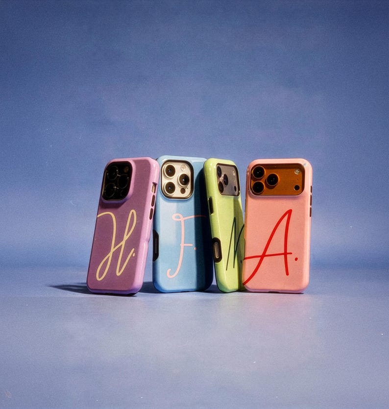 May include: Four colorful phone cases in purple, blue, green, and pink, each with a different handwritten letter. The letters are in a contrasting color to the case. The cases are leaning against a blue background.
