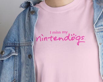 May include: A light pink t-shirt featuring the dark pink text I miss my nintendogs with a small paw print above the letter o. The t-shirt is worn under a blue denim jacket with metal buttons and blue denim jeans, creating a casual layered look.