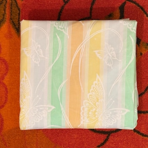 May include: Folded fabric with a striped pattern in pastel colors of yellow, green, and orange. White butterfly and vine designs are printed on the fabric. The fabric is resting on a red floral patterned surface.