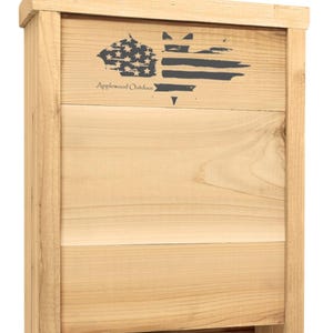 Puede incluir: Una casa para murciélagos de madera con acabado natural. El panel frontal presenta un gráfico gris oscuro de la bandera estadounidense con el texto "Applewood Outdoor". La casa para murciélagos tiene forma rectangular.