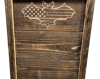 May include: A wooden bat house with a dark brown stained finish. The front panel features a bat silhouette with an engraved American flag design. The bat house is constructed from wooden planks and has a natural wood base.