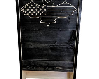 May include: A black wooden bat house with a bat-shaped design featuring the American flag. The bat house is made of wood and has a dark finish. The design is cut out of the wood and painted white.
