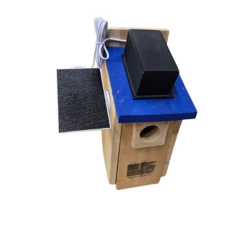 Puede incluir: Una casa para p&aacute;jaros de madera con techo azul y una caja negra encima. Un peque&ntilde;o panel solar est&aacute; adjunto al costado. La casa para p&aacute;jaros tiene un orificio de entrada redondo y una peque&ntilde;a bandera estadounidense.