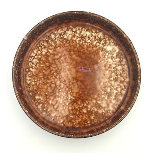 May include: A round, brown ceramic plate with a speckled, textured surface. The plate has a dark brown rim and a lighter brown centre with a speckled pattern. The plate has the text "scheurich" on the bottom.