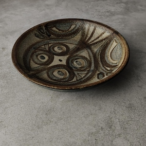 May include: A round, ceramic bowl with a brown and tan color scheme. The bowl features a decorative pattern of swirling lines and circular shapes. The rim is a darker brown, and the bowl has a matte finish. The bowl is likely for serving food or as a decorative piece.