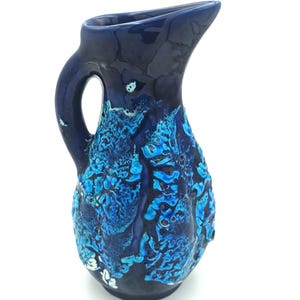 Puede incluir: Una jarra de cerámica azul oscuro con asa y pico vertedor. La superficie de la jarra presenta un diseño abstracto texturizado en tonos azules y blancos. La jarra mide aproximadamente 25 cm de altura.