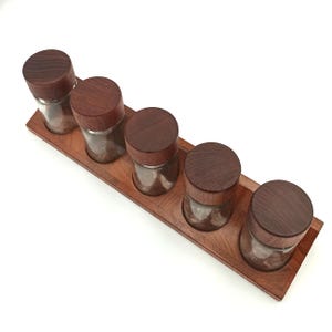 Puede incluir: Un conjunto de cinco botes de especias de vidrio transparente con tapas de madera oscura, dispuestos sobre una bandeja de madera a juego. La bandeja tiene un diseño curvo y es de madera oscura.