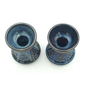 Può includere: Due portacandele in ceramica blu con bordo e base marrone scuro. I portacandele presentano un motivo geometrico sulla base e una finitura liscia e lucida. L'interno dei portacandele è di una tonalità di blu più scura.